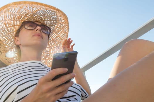 Beautiful, romantic blonde woman on summer vacations traveling by cruse ship ferry boat holding and using mobile phone. Summer vacation lifestyle.