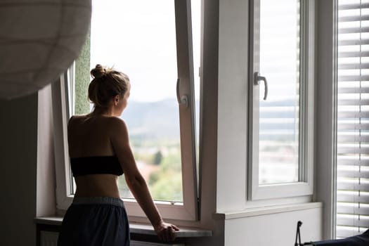 Home quarantine and social distancing during covid pandemic. Silhouette of lonley caucasian woman standing by window, anxiously looking out. Coronavirus infection, pandemics, disease outbreaks.