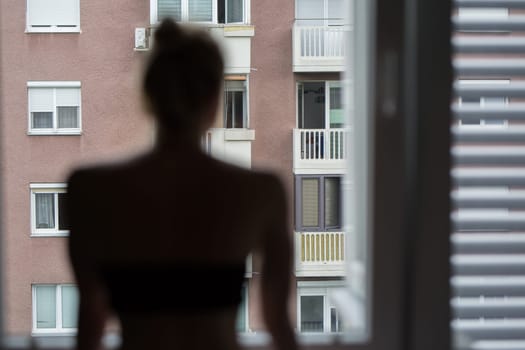 Home quarantine and social distancing during covid pandemic. Silhouette of lonley caucasian woman standing by window, anxiously looking out. Coronavirus infection, pandemics, disease outbreaks.