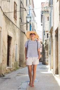 Beautiful blonde young female traveler wearing straw sun hat sightseeing and enjoying summer vacation in an old traditional costal town at Adriatic cost, Croatia.