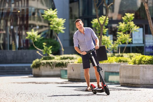 Young casual active sporty businessman in urban city center, wearing shirt and shorts, holding laptop bag, riding to work on electric scooter on a hot summer day.