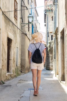 Rear view of beautiful blonde young female traveler wearing straw sun hat sightseeing and enjoying summer vacation in an old traditional costal town at Adriatic cost, Croatia.