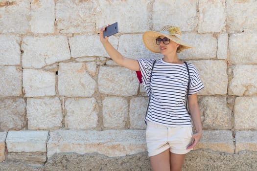 Beautiful young female tourist woman wearing big straw hat, taking self portrait selfie, standing in front of old textured stone wall at old Mediterranean town.