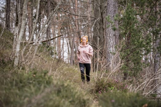 Active sporty woman listening to the music while running in autumn fall forest. Female runner training outdoor. Healthy lifestyle image of young caucasian woman jogging outside.