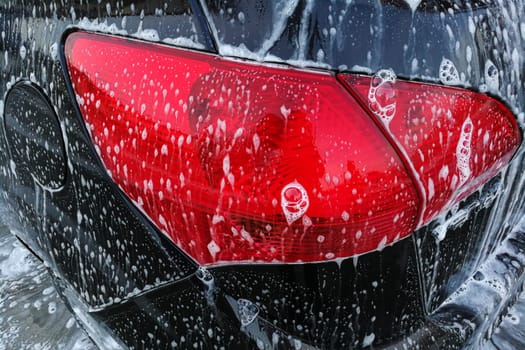 Detail photo of brown car rear red light covered with soap foam when being cleaned in carwash.