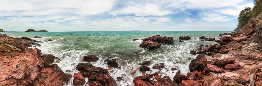High resolution wide panorama of sea waves crushing on red rocks with blue sky with clouds in background. Koh Lanta, Thailand