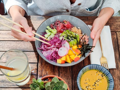 Woman eating tasty colorful healthy natural organic vegetarian Hawaiian poke bowl using asian chopsticks on rustic wooden table. Healthy natural organic eating concept.