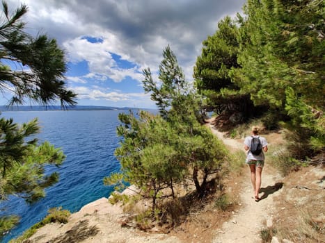 Young active feamle tourist wearing small backpack walking on coastal path among pine trees looking for remote cove to swim alone in peace on seaside in Croatia. Travel and adventure concept.