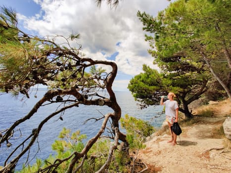 Young active feamle tourist taking a break, drinking water, wearing small backpack while walking on coastal path among pine trees looking for remote cove to swim alone in peace on seaside in Croatia.
