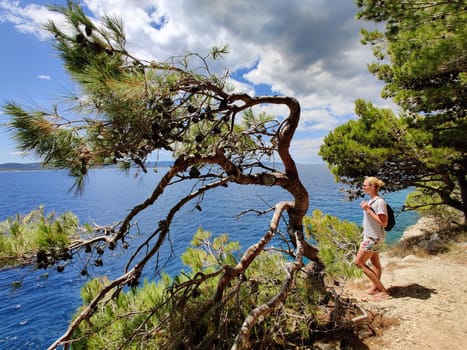 Young active feamle tourist wearing small backpack walking on coastal path among pine trees looking for remote cove to swim alone in peace on seaside in Croatia. Travel and adventure concept.