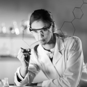 Portrait of a confident female health care expert in life science laboratory writing structural chemical formula on a glass board. Healthcare and modern life science concept. Black and white image.