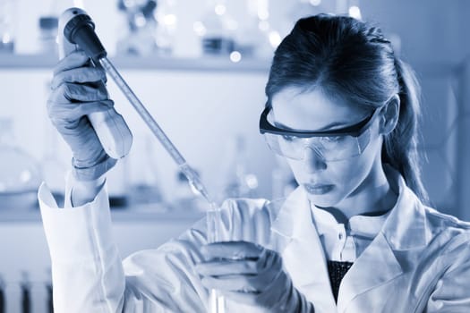 Life scientists researching in laboratory. Focused female life science professional pipetting solution into the glass cuvette. Healthcare and biotechnology concept. Blue toned image.
