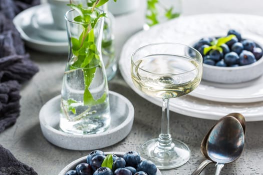 Dining room table decorated with small vase with ivy (Hedera helix). Place setting with plates and bowl with blueberries and glass of wine.