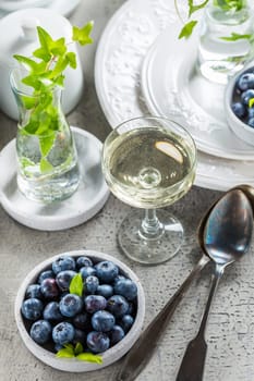 Dining room table decorated with small vase with ivy (Hedera helix). Place setting with plates and bowl with blueberries and glass of wine.