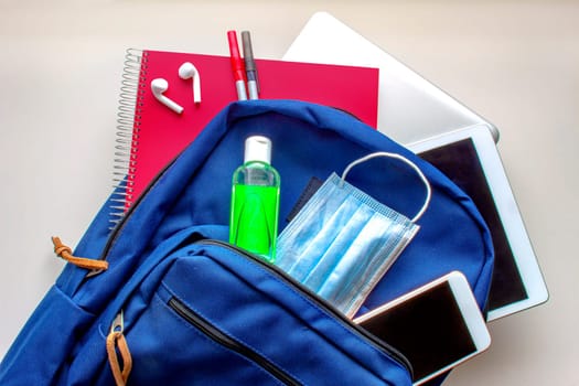 A blue school bag pack with sanitizer and a face mask, pencils earphones, tablet, computer and a smartphone. Back to school during global pandemic. Covid 19.