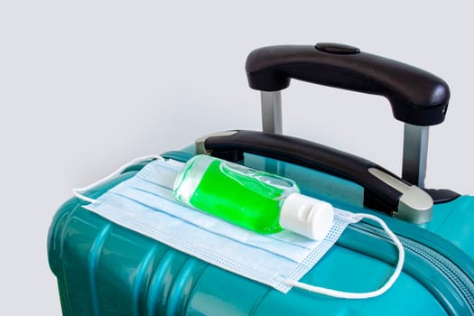 A green luggage with a face mask and a hand sanitizer on a white background. Concept traveling during pandemic. Travel covid 19.