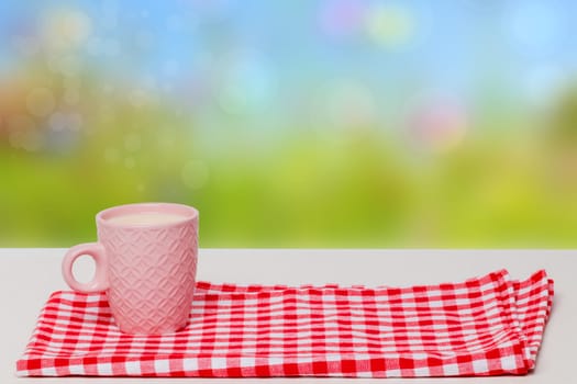 Empty breakfast table. Close-up of a pink ceramic mug with milk on red checkered napkin or tablecloth on a abstract summer background. Template for your food and product display montage.