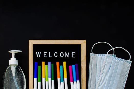 A black board with the text Welcome and markets colors with a hand sanitizer and face masks on a black background