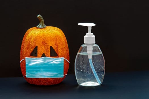 Jack-o-lantern pumpkin Halloween wearing a face mask with hand sanitizer bottle on a black background. Concept: Halloween during global pandemic. Covid-19, Coronavirus.