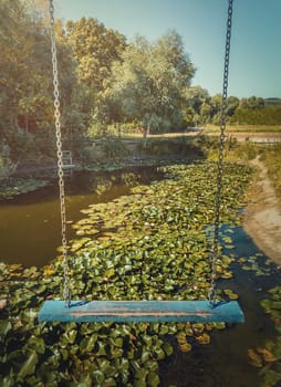 Vertical photo of a vintage blue swing hanging over a pond with blooming waterlily, lotus flowers. Vacation travel, natural background at the lake recreation concept, at Bardar village, Moldova.