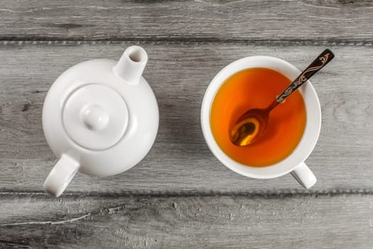 Table top view on small white teapot and cup full of hot amber tea with silver spoon in it, placed on gray wood table.