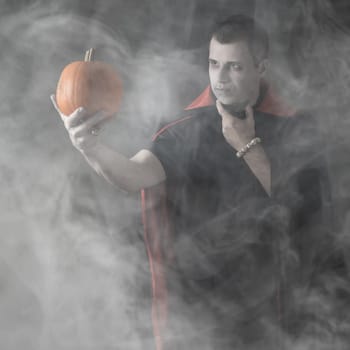 Halloween male vampire portrait. Caucasian white man holding orange pumpkin dressed and stylised for a nosferatu on dark smoky or misty background