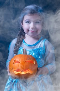 Halloween portrait of young girl stylised for frozen princess in a blue dress. Smiling child holding orange curved pumpkin surrounded by smoke or mist