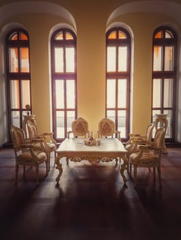 Ancient royalty dining room, medieval furniture style with golden ornate chairs and table near the arched windows. Luxury hosting room background, old wood parquet. Interior architecture details.