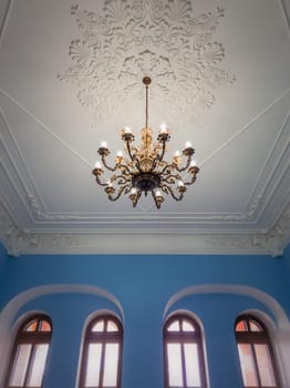 Beautiful golden chandelier suspended down a white ornate ceiling inside a palace room with blue walls and arched windows. Different geometric shapes, ancient interior architecture and design details.