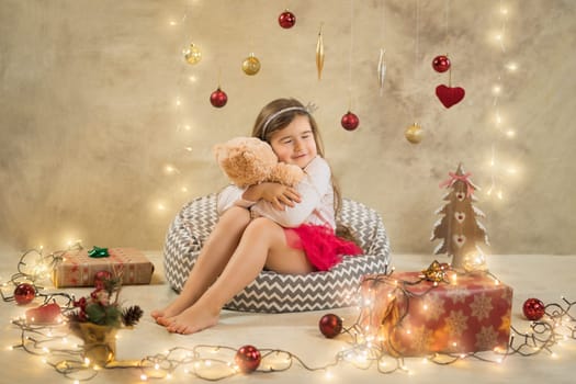 Christmas studio shoot of a cute baby girl that is hugging a teddy bear. Festive, beige background with gifts, Christmas balls and lights