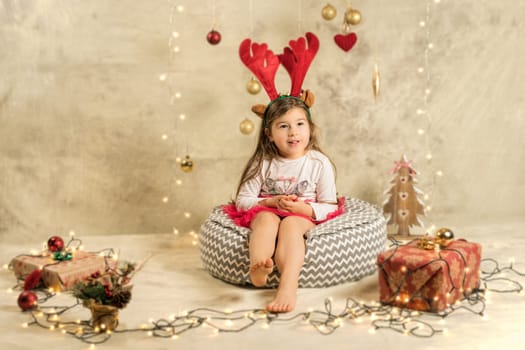 Christmas studio shoot of a cute baby girl wearing reindeer headband. Festive, beige background with gifts, Christmas balls and lights