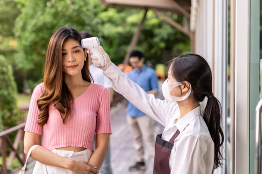 Asian waitress with face mask taking temperature to customer before getting in restaurant with social distance queue in line. New normal restaurant concept after coronavirus covid-19 pandemic.