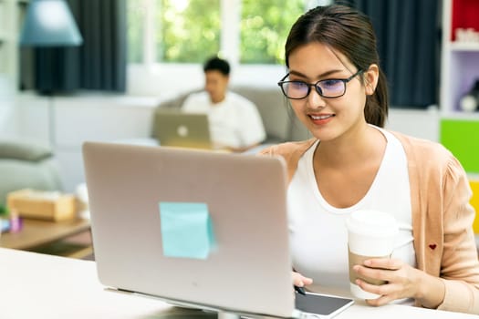 Asian working woman use computer notebook laptop with delivery coffee cup work from home in living room with social distance to couple in background while city lockdown. Work at home concept.