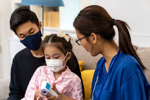 Asian female doctor show temperature using infrared thermometer to little girl child sit with her dad in living room while doctor visit at home. Home health care delivery and doctor visiting concept.