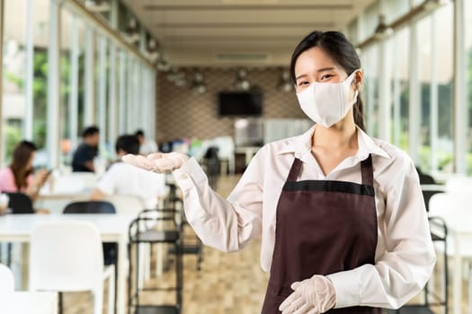Portrait attractive asian waitress wear face with background of customers sit in social distance table in restaurant indoor. New normal restaurant lifestyle concept.