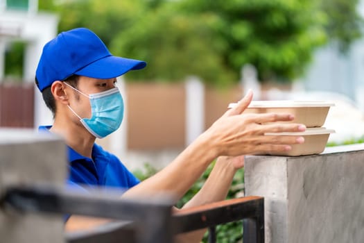 Asian deliver man with face mask in blue shirt handling food boxes and put in on the fence post of customer house as contactless food delivery. This can reduce coronavirus COVID-19 spreading.