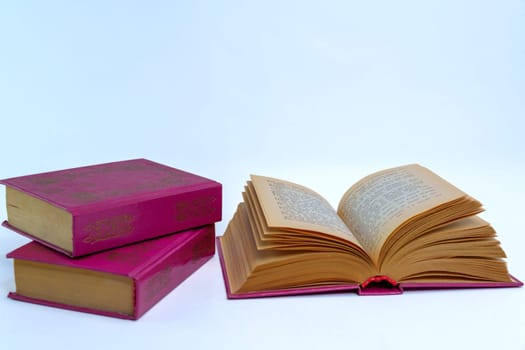 An open vintage hardcover book with yellowed pages next to a stack of two closed books on a neutral background. Classic literature concept representing reading, knowledge, and education