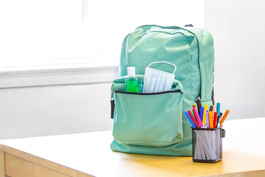 A student school bag pack with sanitizer and a face mask and pencil markers on a table. Back to school during global pandemic. Covid 19.