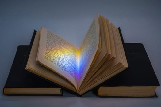 An open vintage book with a colorful light reflection on its pages, resting on two stacked black hardcover books. Symbolic concept of knowledge, mystery, and inspiration