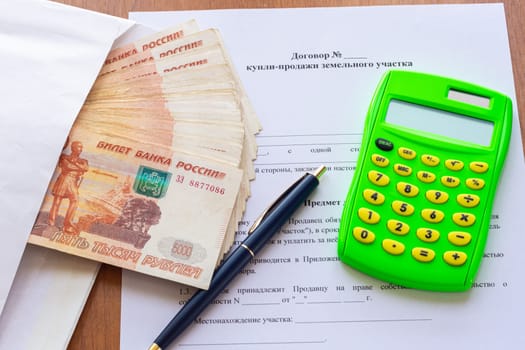 On the table is a contract for the sale of a land plot, a bundle of money in an envelope, a pen and a calculator