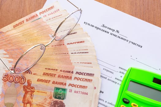 On the table there is a contract for the sale of a land plot, a bundle of money, a calculator and glasses
