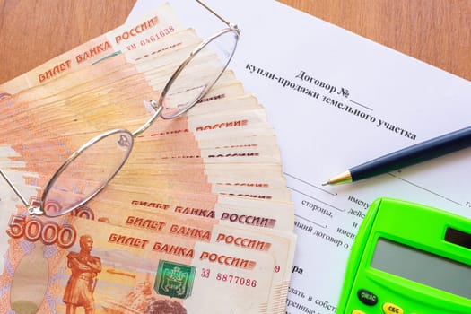 On the table is a contract for the sale of a land plot, a bundle of money, a calculator, glasses and a pen