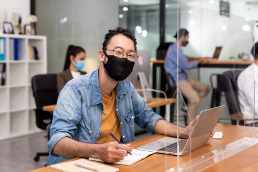 Portrait of asian office employee businessman wear protective face mask work in new normal office with interracial colleague in background as social distance practice prevent coronavirus COVID-19.