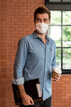Portrait of middle east caucasian businessman with takeout coffee cup and clipboard in meeting room after lunch break. He wear face mask as new normal office preventing coronavirus COVID-19 spreading.
