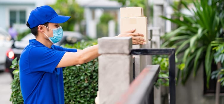 Panoramic side view asian deliver man with face mask in blue shirt handling packages and put in on the fence post as contactless shopping delivery. This can reduce coronavirus COVID-19 spreading.