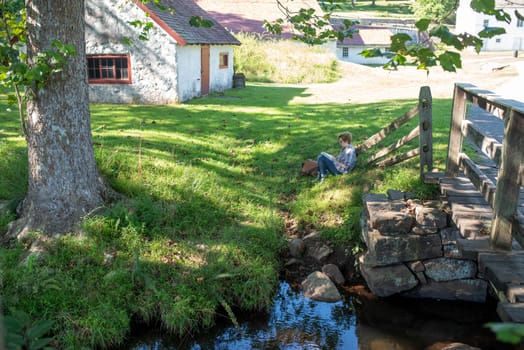 Tranquil early American villiage scene at Hopewell Furnace National Historic Site in Pennsylvania. Young woman relaxes alone in the shade reading a book.