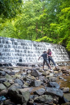 Water cascades over the tiered waterfall with lush woodland foliage behind. Vertical image of two young men, African-American and Caucasian, at the base of falls.