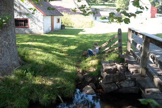 Tranquil early American villiage scene at Hopewell Furnace National Historic Site in Pennsylvania. Young woman relaxes alone under a tree by a stream reading a book.