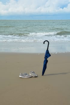 A closed umbrella is stuck in the sand and old shoes with holes stand on the beach in front of the sea with many waves