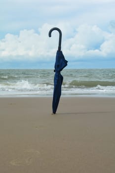 A closed umbrella is stuck in the sand on the beach in front of the ocean with waves, sky and copy space